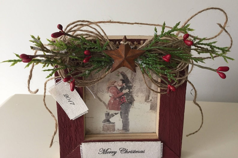 May include: A rustic red wooden picture frame decorated with greenery, red berries, and a rusty star. The frame holds a black and white photo of a couple kissing. The text "Merry Christmas" is at the bottom, and a tag reads "2017".