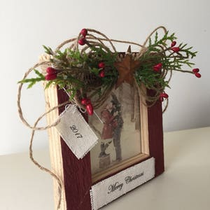 May include: A rustic wooden picture frame with a photo of a couple in the snow. The frame is painted burgundy and decorated with twine, greenery, red berries, and a star. The frame has a tag that says "2017" and a banner that says "Merry Christmas".