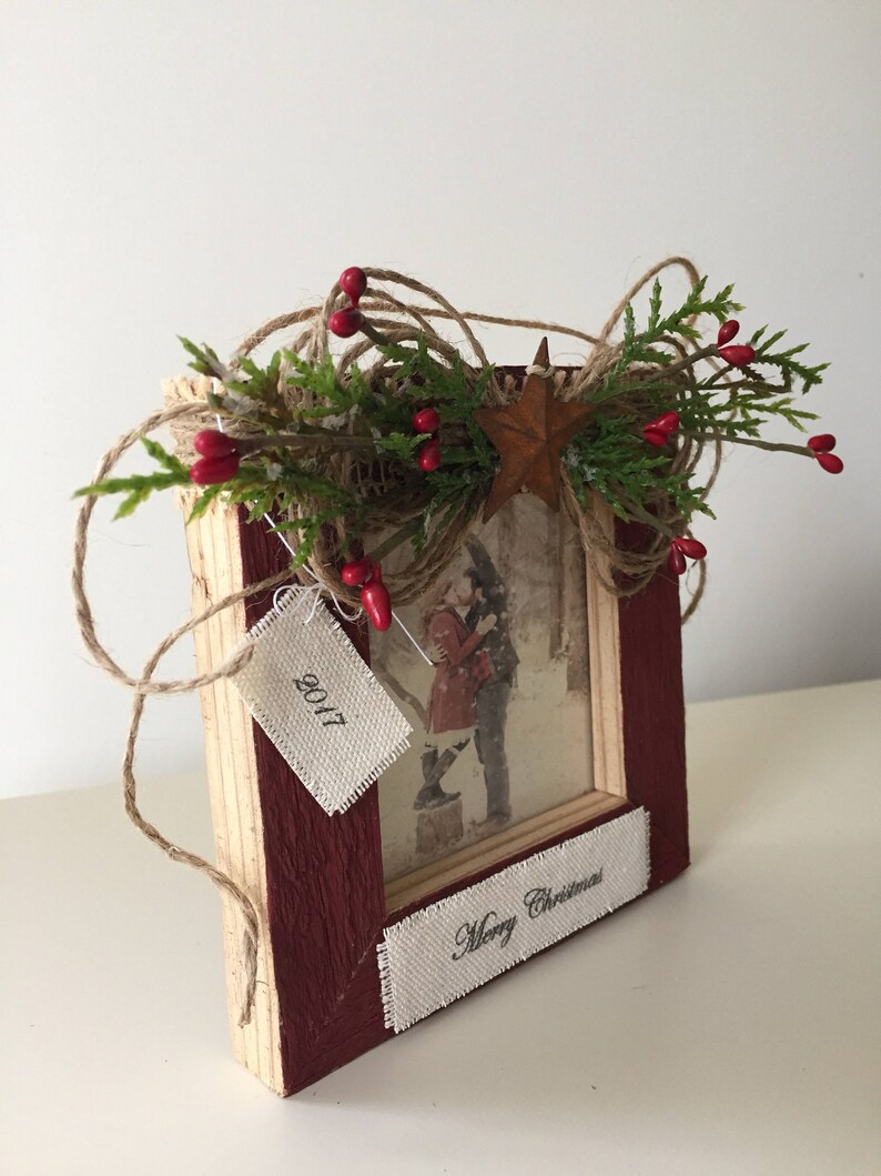 May include: A rustic wooden picture frame with a photo of a couple in the snow. The frame is painted burgundy and decorated with twine, greenery, red berries, and a star. The frame has a tag that says "2017" and a banner that says "Merry Christmas".