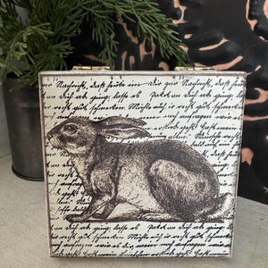 May include: A small, rectangular wooden box with a hinged lid. The box features a black and white illustration of a rabbit and script-like text. The box is likely used for storage or decoration.