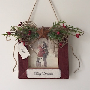 May include: A framed Christmas decoration with a rustic aesthetic. The burgundy frame holds a winter scene print and a banner reading "Merry Christmas." Embellishments include a star, twine, greenery, and red berries. A tag reads "2017."