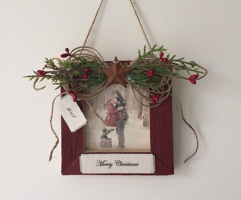 May include: A framed Christmas decoration with a rustic aesthetic. The burgundy frame holds a winter scene print and a banner reading "Merry Christmas." Embellishments include a star, twine, greenery, and red berries. A tag reads "2017."