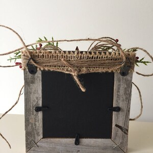 May include: A rustic, square picture frame with a weathered, gray wooden frame. The top is decorated with burlap, twine, green sprigs, and red berries. The frame has a black backing and metal hardware.