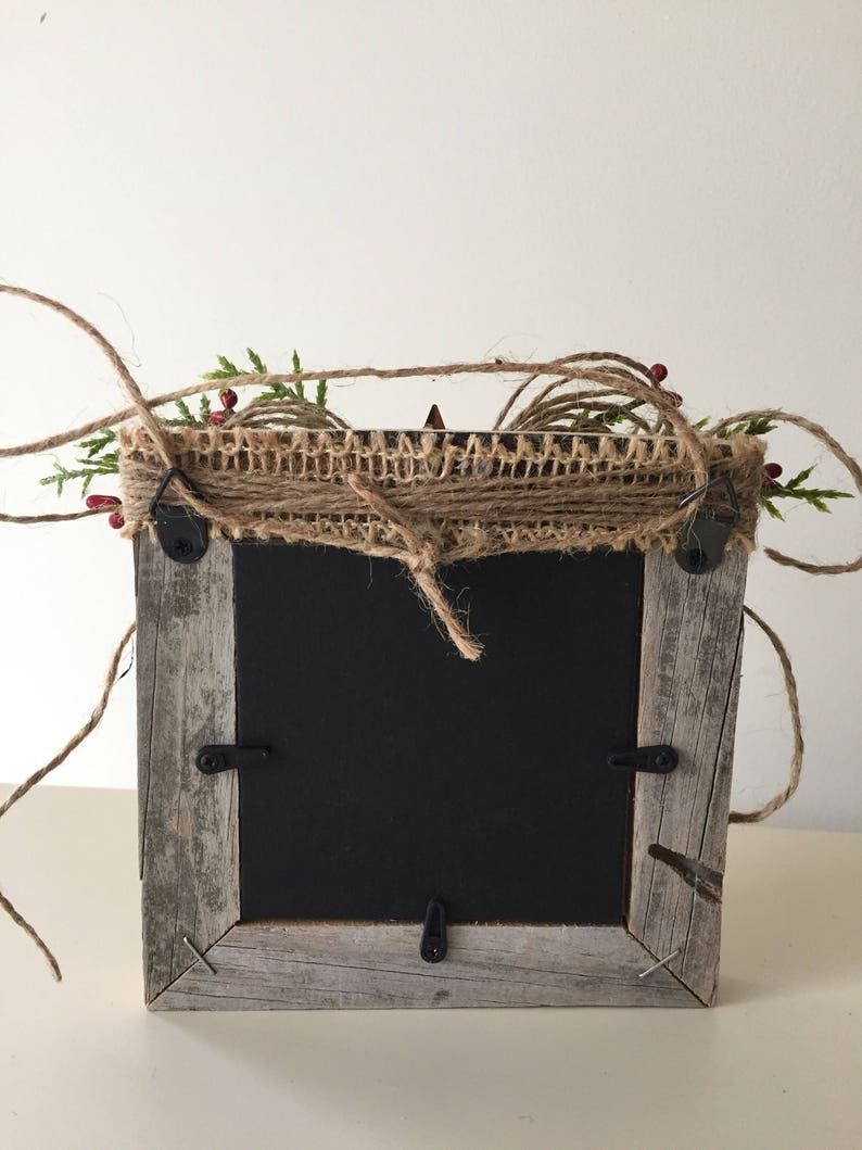 May include: A rustic, square picture frame with a weathered, gray wooden frame. The top is decorated with burlap, twine, green sprigs, and red berries. The frame has a black backing and metal hardware.