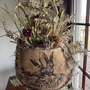 May include: A vintage Easter decoration featuring a paper container with a black and white illustration of two rabbits and the words "Happy Easter." The container is filled with dried flowers and greenery, and trimmed with silver tinsel.