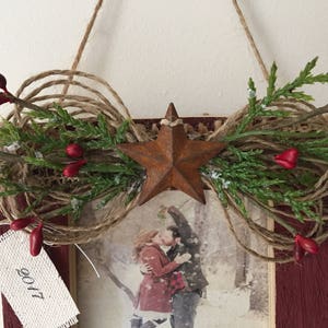 May include: Rustic wooden framed picture with a star and twine embellishment. The frame is decorated with greenery, red berries, and a rusty star. A tag with "2017" hangs from the frame. The picture inside shows a couple kissing.