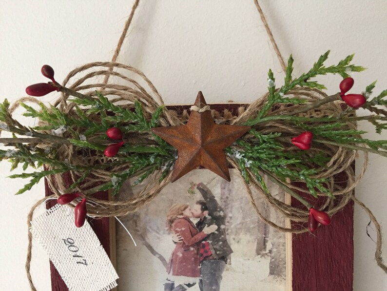 May include: Rustic wooden framed picture with a star and twine embellishment. The frame is decorated with greenery, red berries, and a rusty star. A tag with "2017" hangs from the frame. The picture inside shows a couple kissing.