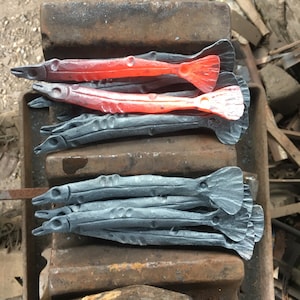 May include: Hand-forged metal fish sculptures in various stages of completion. Two are glowing red, while others are dark grey. The fish are lying on a rusty metal anvil, showcasing the blacksmithing process.
