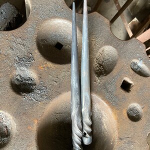 May include: Two hand-forged metal spikes with tapered points and twisted handles. Each spike features a circular hole at the base of the handle. The metal has a dark, textured, matte grey finish consistent with ironwork, showing signs of forging.