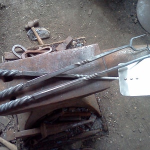 May include: A set of three forged metal fireplace tools, including a poker, tongs, and a shovel. The tools have twisted handles and are resting on an anvil.