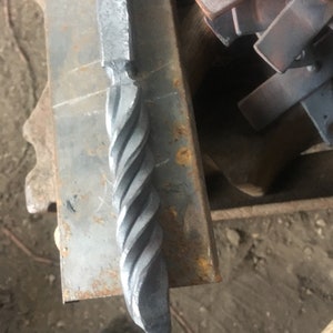 May include: Close-up of a hand-forged metal object with a twisted, spiral design. The metal has a rough, textured surface and a dark gray color. The object appears to be a decorative element or part of a larger piece.