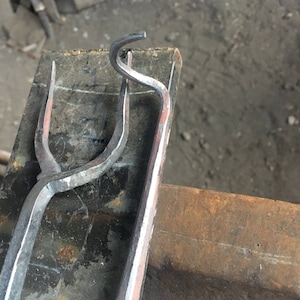 May include: Close-up of a metal hook being forged on an anvil. The hook is bent into a U-shape with a pointed end.