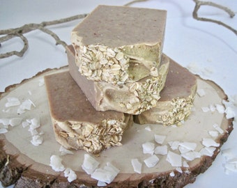 May include: A stack of four brown and beige soap bars with oatmeal on top, sitting on a wooden slice with white coconut flakes scattered around.