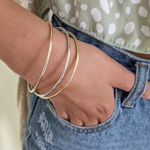 Può includere: Un set di tre braccialetti rigidi dorati e argentati impilati sul polso di una persona. I braccialetti sono sottili e delicati, e sono un accessorio di moda popolare.