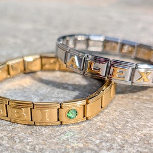 May include: Two personalized charm bracelets. One is gold-toned with the name "BIBI" and a green gem. The other is silver-toned with gold accents and the name "ALEX". Both bracelets are linked with rectangular segments.