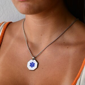 May include: A silver chain necklace with a silver octagonal pendant featuring a blue medical symbol.