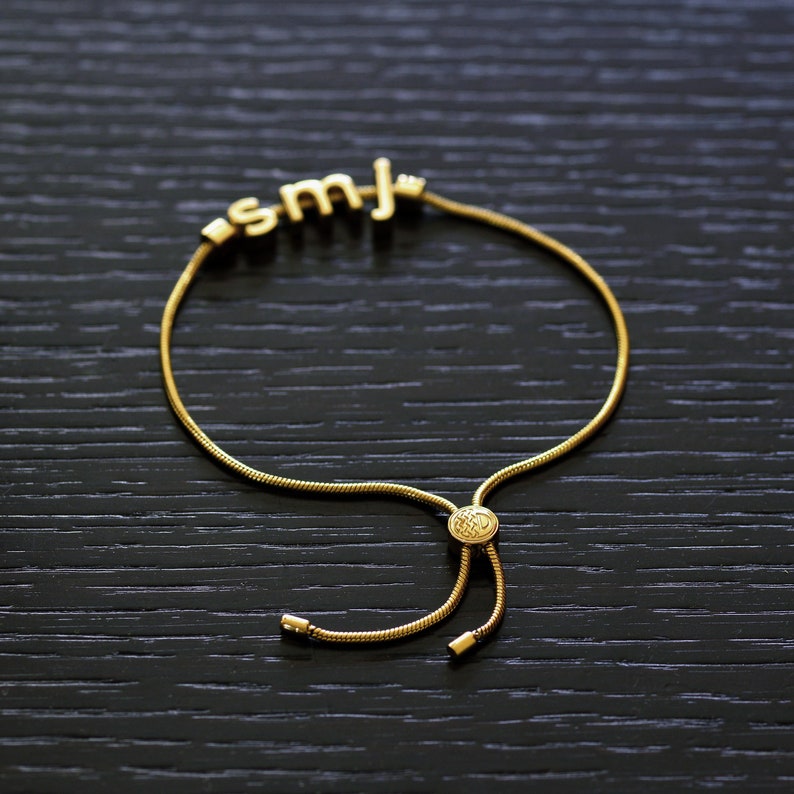 K&ouml;nnte beinhalten: Ein goldenes Kettenarmband mit einem Schiebeverschluss und einem Anh&auml;nger mit den Buchstaben "SMJ".