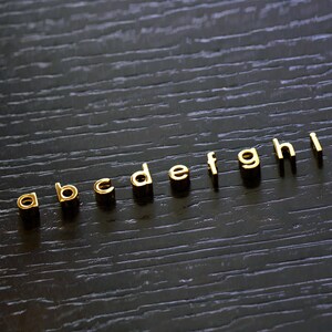 K&ouml;nnte beinhalten: Goldfarbene Metallbuchstaben, die das Alphabet von "a" bis "i" auf einer dunklen Holzoberfl&auml;che buchstabieren.