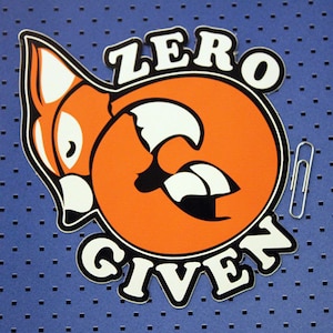 Peut inclure: Un autocollant noir et orange avec le texte "ZERO GIVEN" en contour blanc. L'autocollant représente un renard de dessin animé avec un visage blanc et un corps orange.