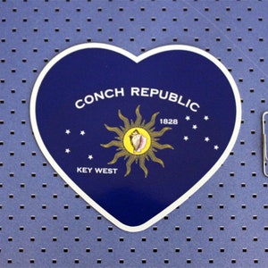 May include: A blue heart-shaped sticker with the text "Conch Republic" and "Key West" in white. A yellow sun with a pink conch shell is in the center. The year "1828" is printed above the shell.