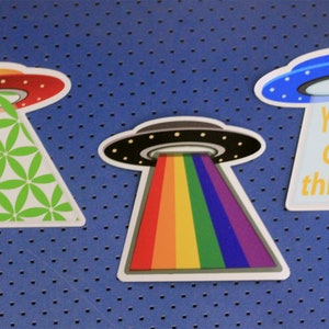 May include: Three colorful UFO stickers with different designs. The first sticker features a green and white geometric pattern. The second sticker has a rainbow beam of light. The third sticker is blue and white with the text "You're outta this world".