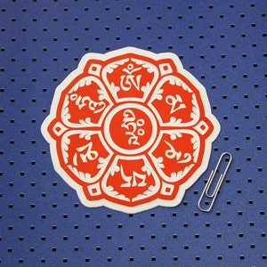 May include: A white sticker with a red lotus flower design. The flower has eight petals with a white center. Each petal has a different Tibetan symbol in white.