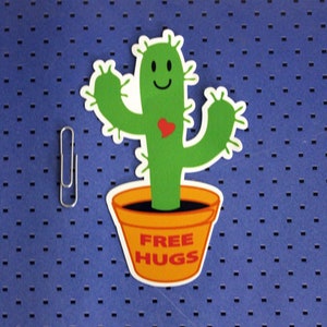 May include: A green cactus with a smiling face and a red heart on its body is in a yellow pot with the words "FREE HUGS" on it.
