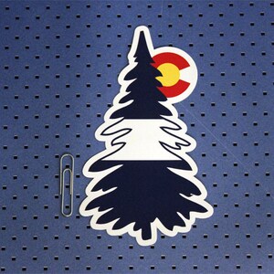 May include: A white paper sticker with a black silhouette of a pine tree. The tree is divided into three sections: a dark blue section at the bottom, a white section in the middle, and a dark blue section at the top. The top section has a red circle with a yellow circle inside, representing the Colorado state flag.