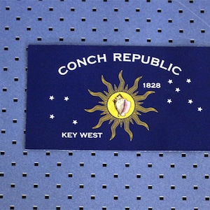 May include: A blue flag with the words "Conch Republic 1828" and "Key West" printed in white. A yellow sun with a conch shell in the center is depicted on the flag.