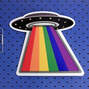 May include: A rainbow-colored beam of light shoots out from a flying saucer, which is a paper cutout on a blue background.