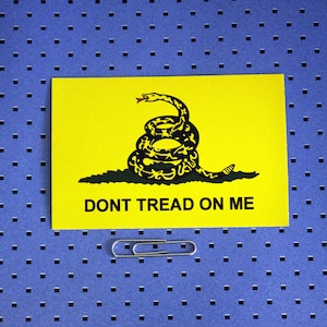 May include: A yellow flag with a black rattlesnake coiled and ready to strike. The snake has a red tongue and the words "DONT TREAD ON ME" are printed in black below the snake.