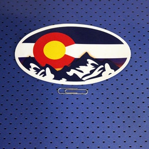 May include: Oval sticker with a graphic of the Colorado state flag and mountains. The flag is red, yellow, and white with a blue background. The mountains are white with a blue background.
