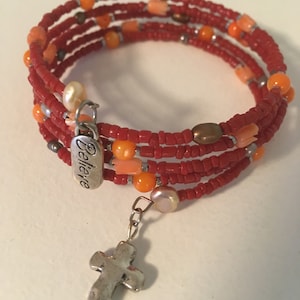 Puede incluir: Una pulsera envolvente con cuentas de semillas rojas, cuentas naranjas y perlas de agua dulce. Un dije plateado con la palabra "Believe" y un colgante de cruz plateada cuelgan de la pulsera. El brazalete es de diseño multi-hebra.