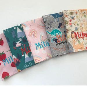 May include: Five colorful fabric napkins with various patterns. One features strawberries, another mountains, and a third rainbows. The fourth has dinosaurs, and the last one has woodland creatures. Each napkin has the name "Mila" embroidered on it.