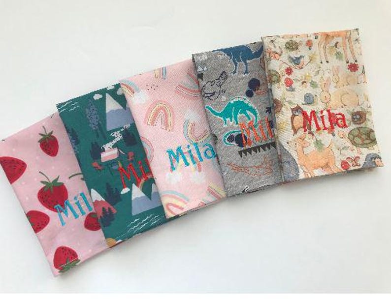 May include: Five colorful fabric napkins with various patterns. One features strawberries, another mountains, and a third rainbows. The fourth has dinosaurs, and the last one has woodland creatures. Each napkin has the name "Mila" embroidered on it.