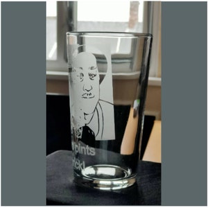 Puede incluir: Un vaso de pinta de vidrio transparente con un dise&ntilde;o grabado en blanco de la cara de un hombre y el texto "pints nickl".