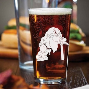 May include: A clear glass pint glass with a white design of a cartoon dwarf holding a pickaxe. The glass is filled with a dark amber colored liquid.