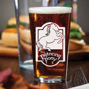 May include: A clear glass pint glass filled with brown beer. The glass features a white design of a horse and the text "The Prancing Pony".
