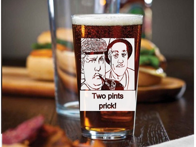 Puede incluir: Un vaso de pinta de vidrio transparente con cerveza marr&oacute;n en su interior. El vaso tiene una imagen en blanco y negro de dos hombres con el texto "Two pints prick!" impreso en la parte delantera.