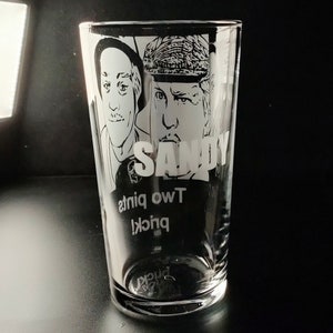 Puede incluir: Un vaso de pinta de vidrio transparente con un dise&ntilde;o grabado en blanco y negro de dos caras de hombres y el texto "SANDY" y "TWO pints" en la parte inferior.