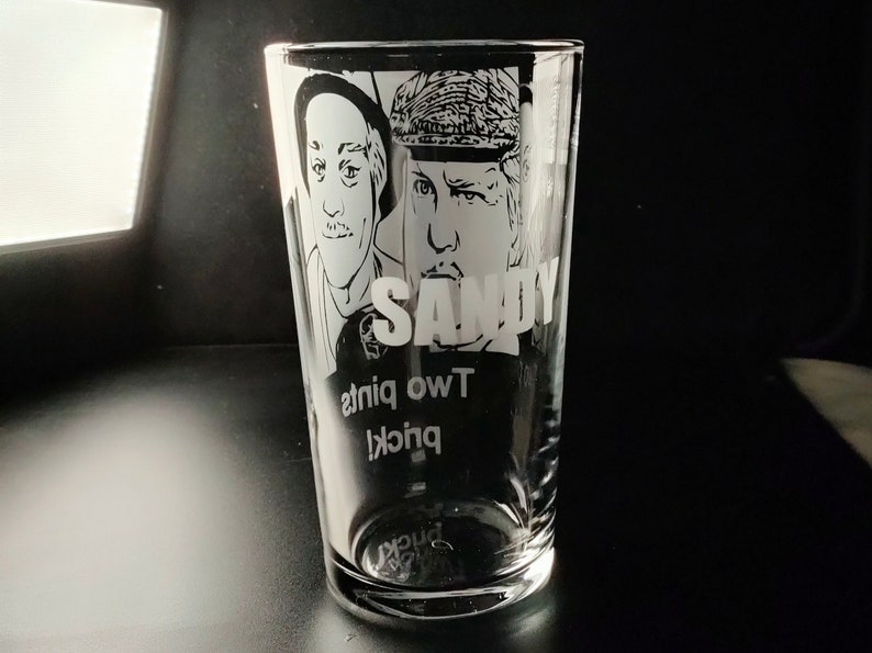 Puede incluir: Un vaso de pinta de vidrio transparente con un dise&ntilde;o grabado en blanco y negro de dos caras de hombres y el texto "SANDY" y "TWO pints" en la parte inferior.