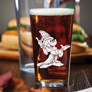 May include: A clear glass pint glass filled with brown beer. The glass features a white silhouette of Mickey Mouse dressed as a sorcerer.