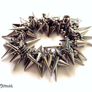 Op de afbeelding: Een zilveren armband met een cirkel van scherpe metalen spikes. De armband is gemaakt van metalen kettingschakels en heeft een punkrock-esthetiek.