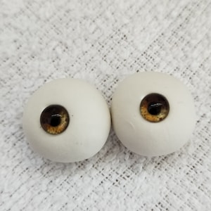 May include: A pair of doll eyes, each with a white, spherical outer shell and a dark brown iris with gold and brown detailing. They are placed on a white, textured surface.
