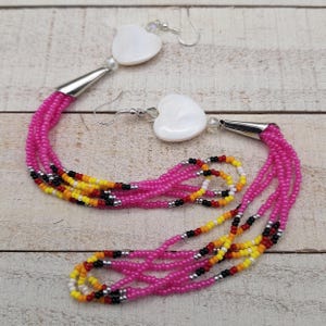May include: A pair of dangle earrings featuring white heart-shaped pendants and multiple strands of pink beads. The beads transition to a pattern of yellow, orange, black, and white beads. Silver-toned metal accents the design.