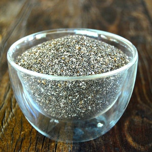 Könnte beinhalten: Eine Glasschale gefüllt mit Chia-Samen. Die Samen sind klein, ovalförmig und variieren in der Farbe von Schwarz über Grau bis Weiß. Die Schale steht auf einer dunklen, strukturierten Holzoberfläche.