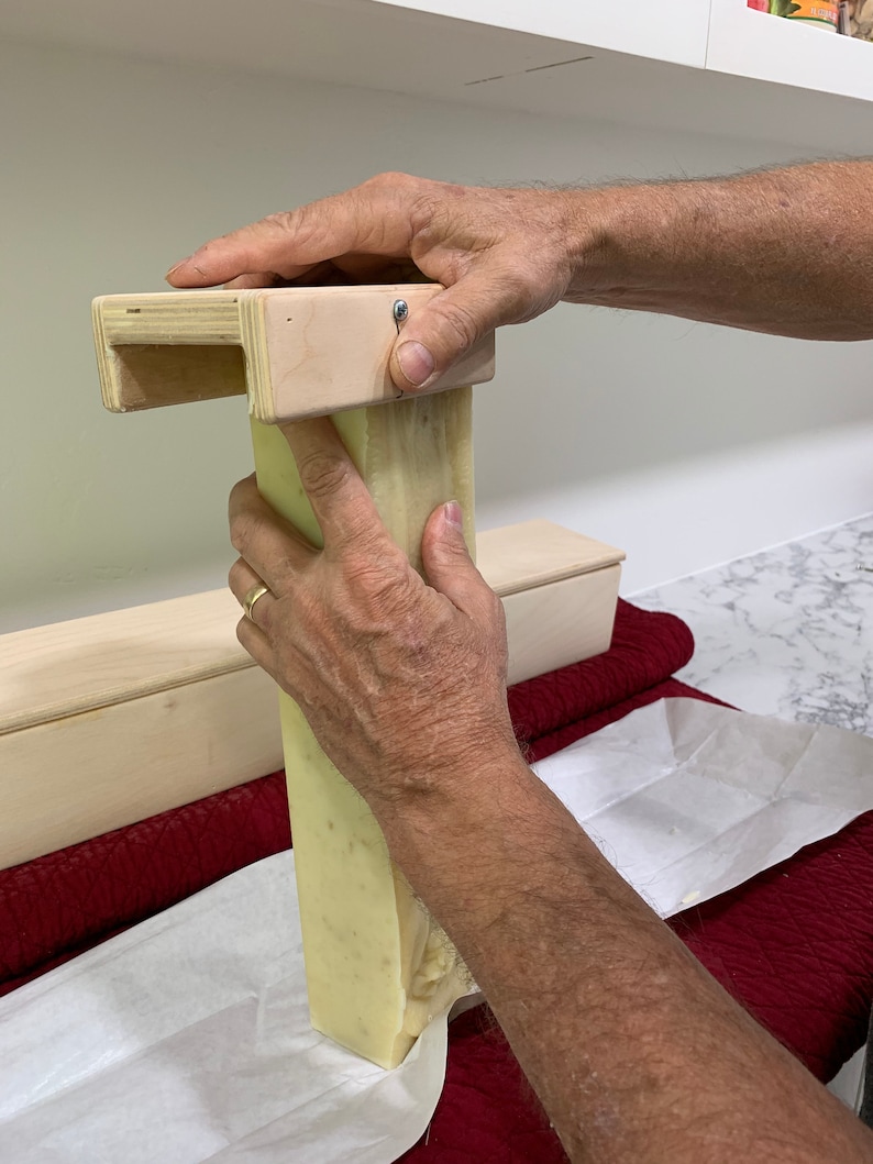K&ouml;nnte beinhalten: Die H&auml;nde einer Person dr&uuml;cken auf eine h&ouml;lzerne Seifenform. Ein St&uuml;ck hellgr&uuml;ner Seife wird aus der Form gedr&uuml;ckt. Die Seife liegt auf einem wei&szlig;en Blatt Papier.