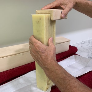 K&ouml;nnte beinhalten: Eine Person verwendet einen h&ouml;lzernen Seifencutter, um einen gro&szlig;en Block selbstgemachter Seife zu schneiden. Die Seife ist hellgelb und hat eine k&ouml;rnige Textur. Die Seife wird auf einem wei&szlig;en Blatt Papier geschnitten.
