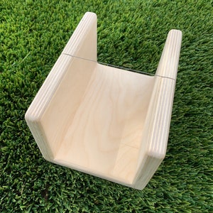 May include: A wooden ramp with a natural finish, featuring a smooth, angled surface and vertical side supports. The ramp is designed with a simple, geometric shape and is set against a backdrop of green artificial turf.