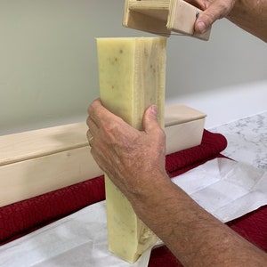 K&ouml;nnte beinhalten: Eine Person schneidet einen gro&szlig;en Block selbstgemachter Seife mit einem h&ouml;lzernen Seifenschneider. Die Seife ist hellgelb und hat eine k&ouml;rnige Textur. Die Seife liegt auf einem wei&szlig;en Blatt Papier.